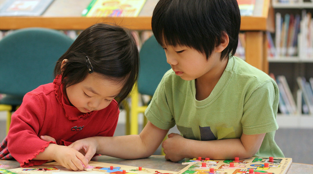A pair toddlers work on a wooden puzzle together in a library.