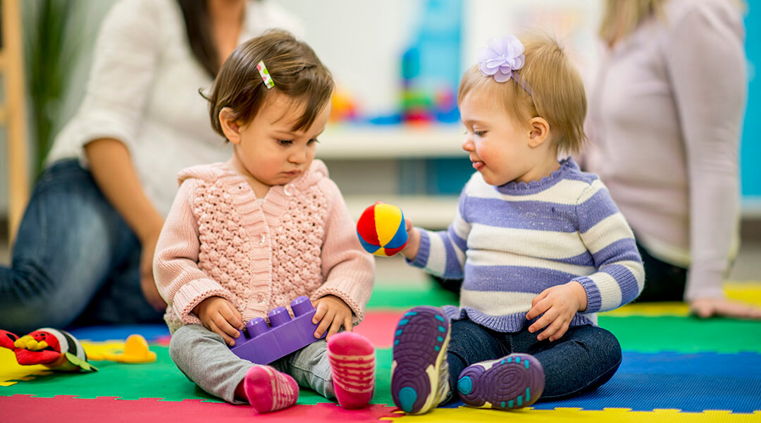 A baby shares a ball with her friend; their caregivers chat in the background.