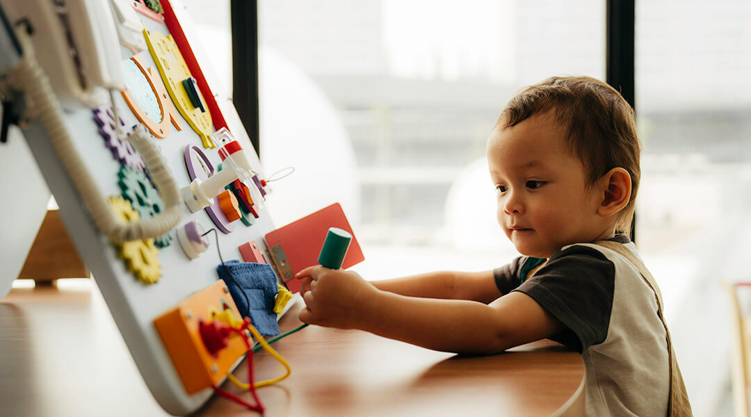 A toddler plays with a busy board which has gadgets like gears, cords, and handles.