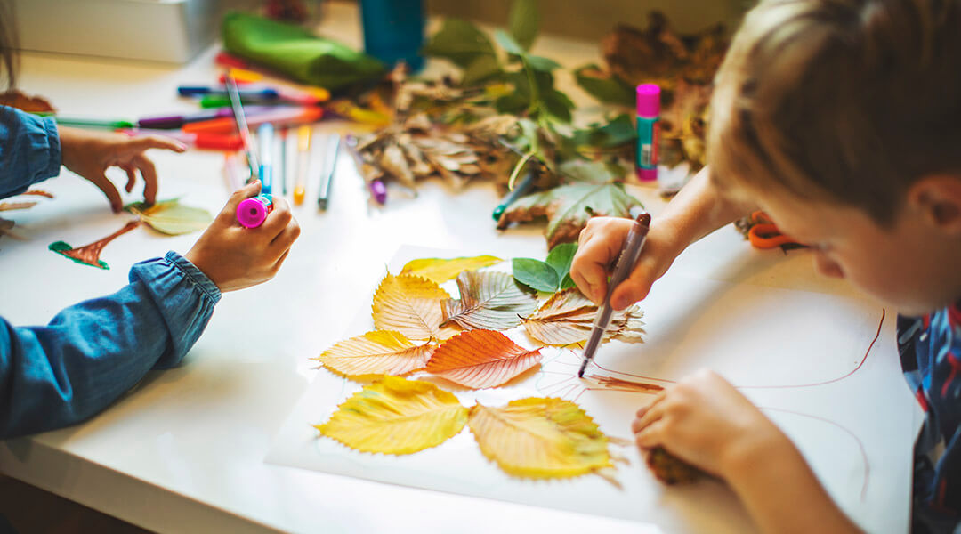 Two children do crafts with leaves and markers.