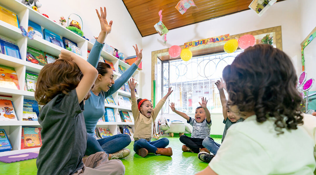 A group of pre-schoolers sit on the floor with their hands in the air during a circle time.