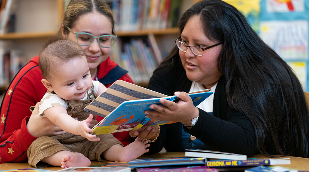 Two adult caregivers reading a board book to a baby