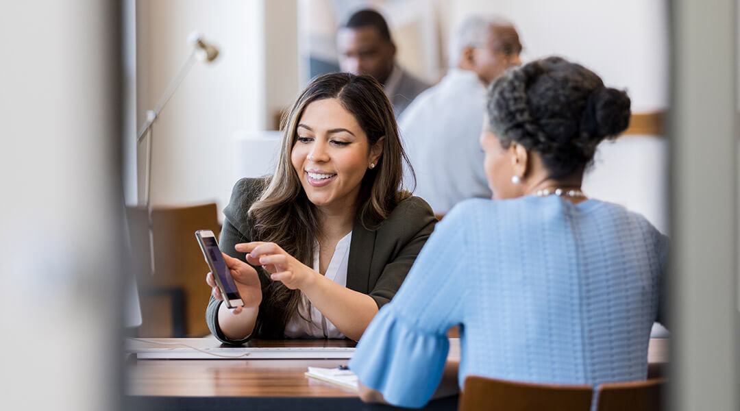 An adult shows their phone screen to another person, pointing out a specific feature.