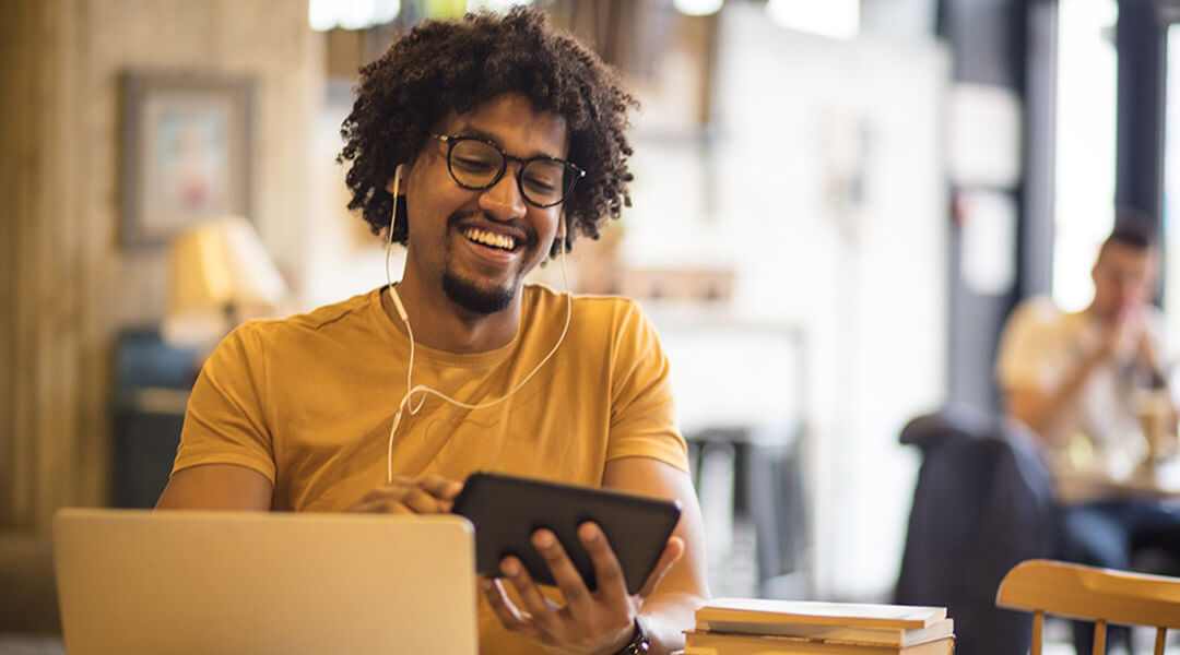 A person wearing headphones looks at their phone with a laptop open in front of them.