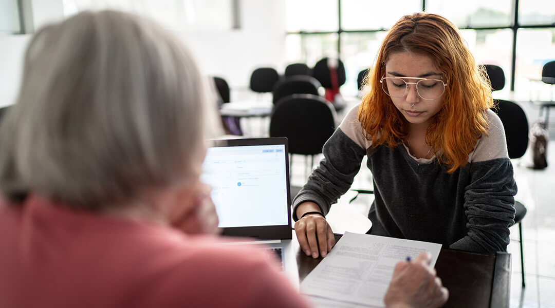 A person at a desk reviews the paperwork of the young adult meeting with them.