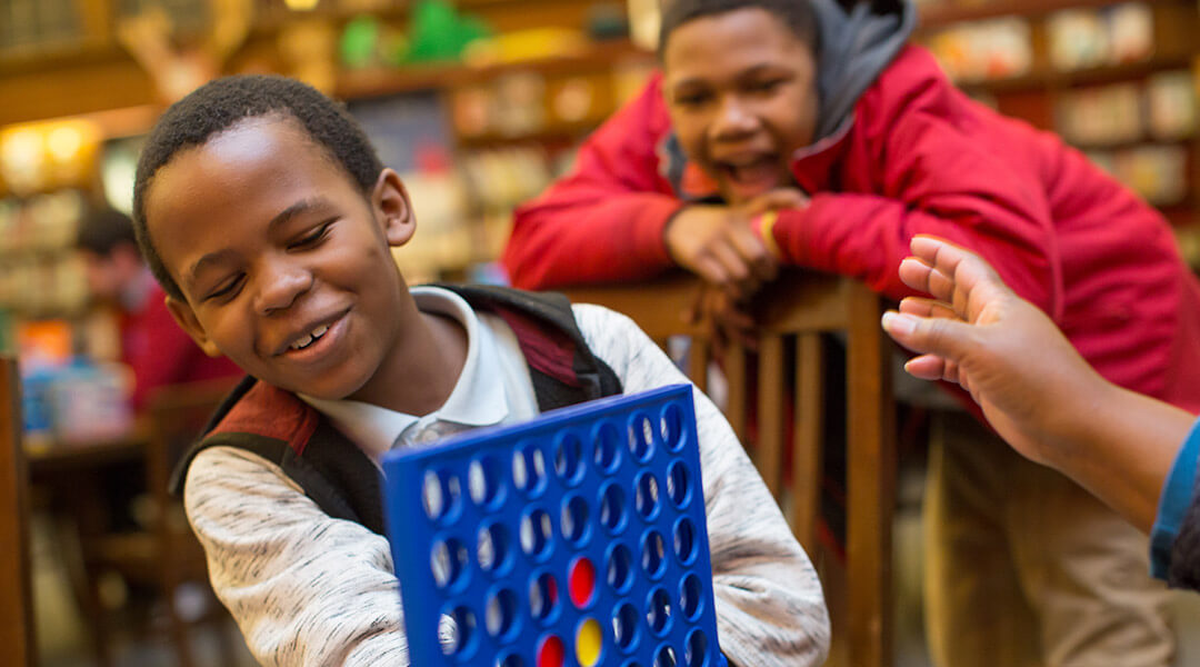 A child plays connect four while another watches in the background, smiling.