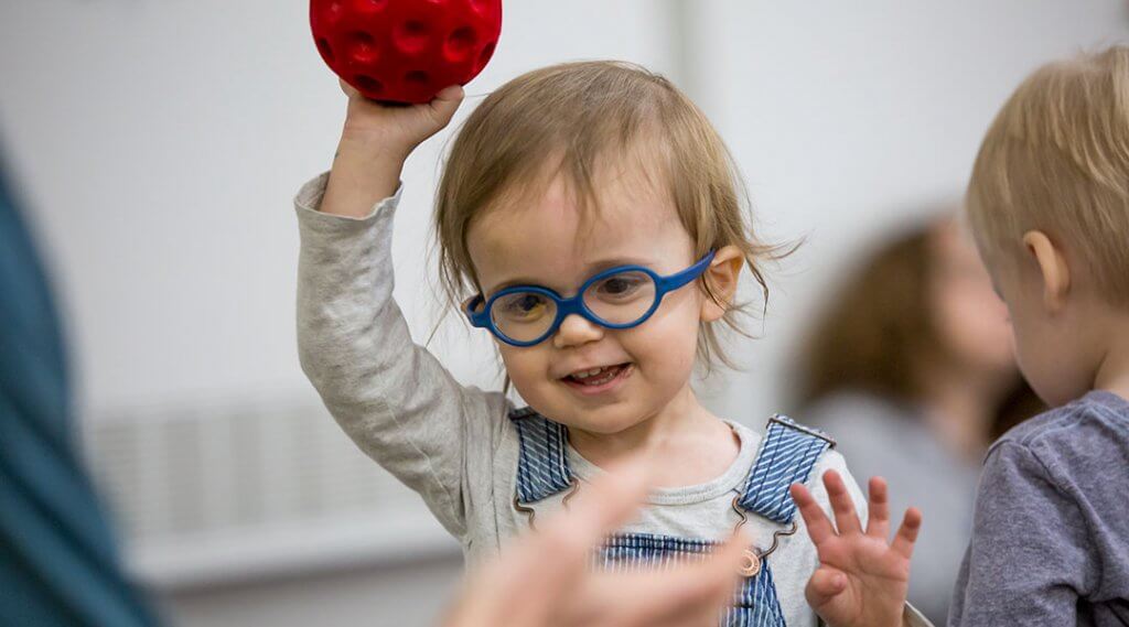 A toddler wearing bright blue glasses holds a ball in the air.