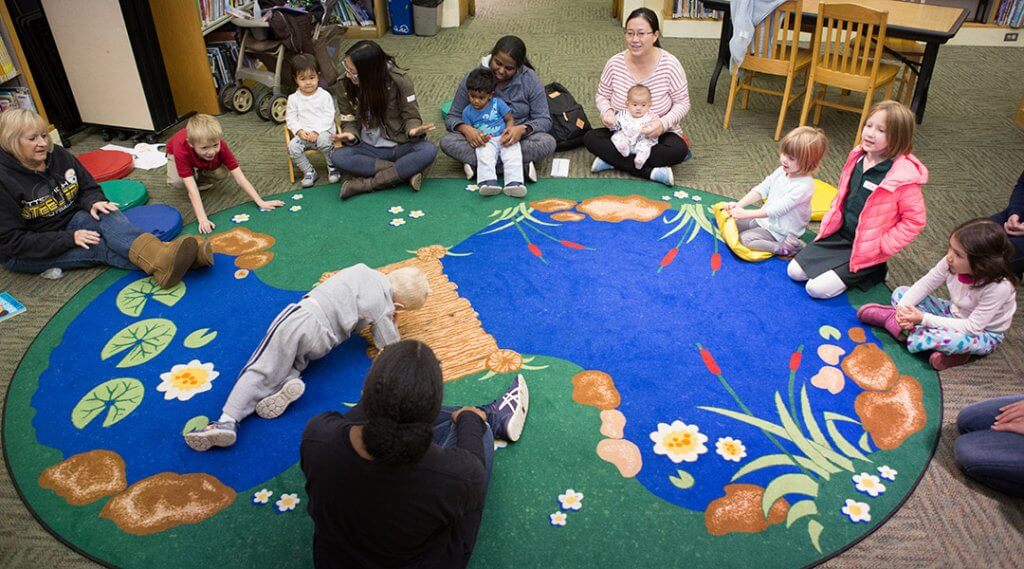 Small children and their caregivers sit in a circle around a carpet that looks like a pond, ready for storytime.