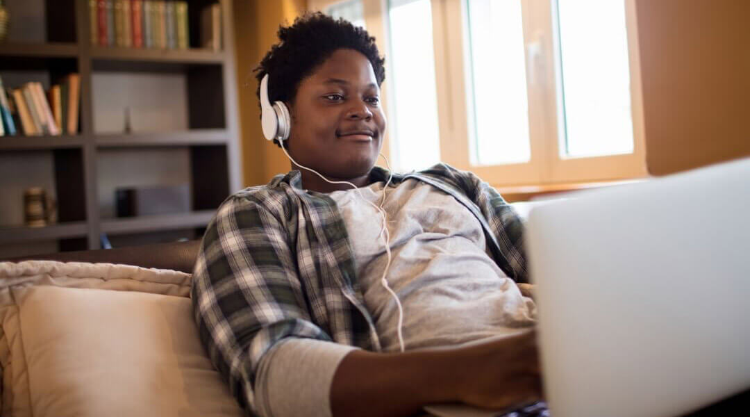 Teen reclining on a couch while wearing headphones and using a laptop.