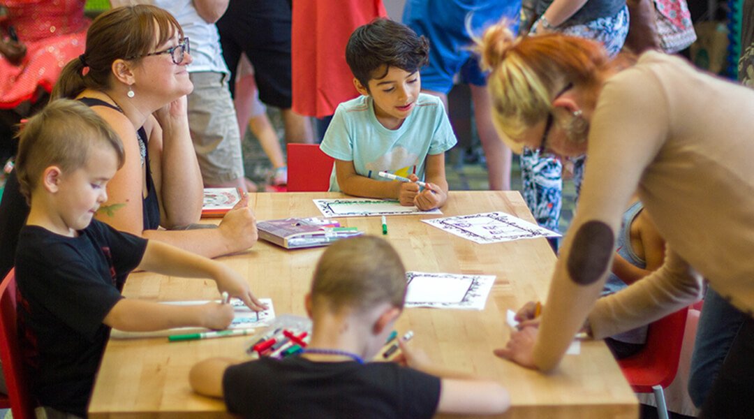 A few young children share a table with adults as they all color with markers.
