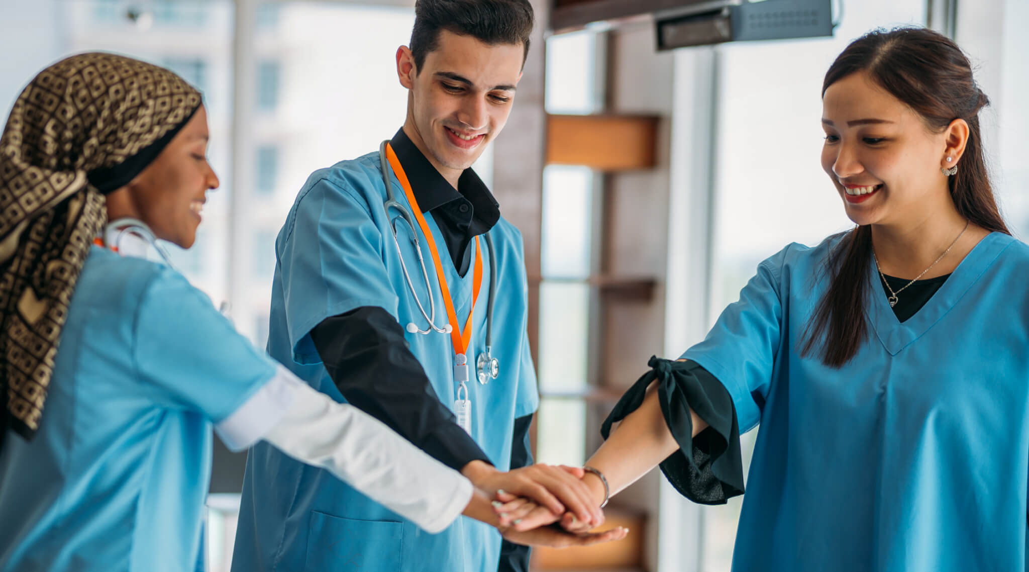 Three nurses wearing scrubs layer their hands in preparation for a 'go team' type motion.