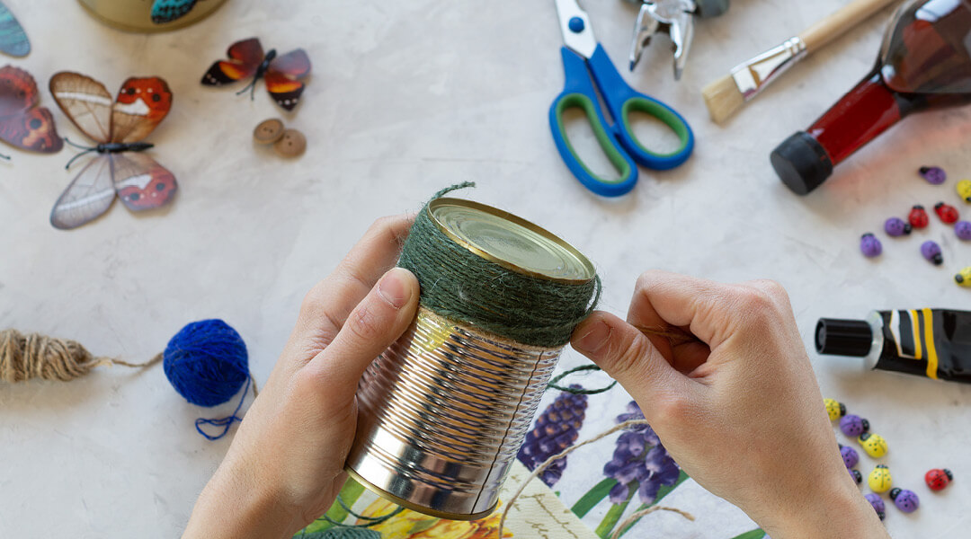 Close-up of hands wrapping green twine around a tin can with other craft supplies scattered in the background.