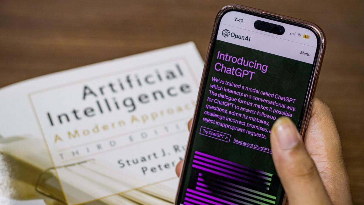 A hand holding a phone with text visible that reads "OpenAI: Introducing ChatGPT." In the background, the book "Artificial Intelligence: A Modern Approach" by Stuart J. Russell and Peter Norvig sits atop a table, slightly out of focus.