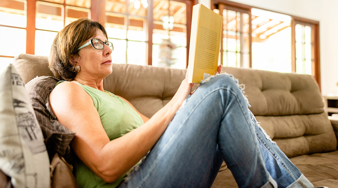 Mature adult spending a relaxing afternoon reading a book on their living room sofa at home