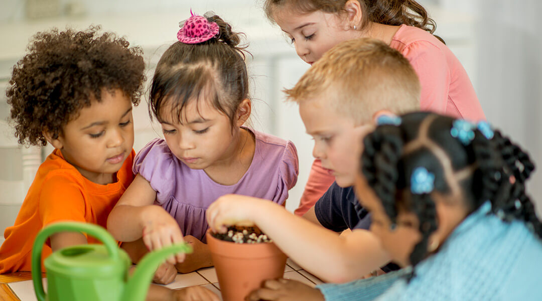 Five young children plant seeds in a small clay pot.