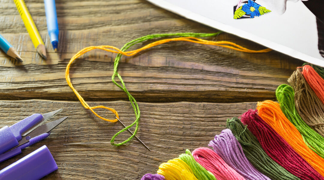 Close-up of a needle and multi-colored thread on a table, connected to a piece of paper with embroidered stitches to look like a flower.