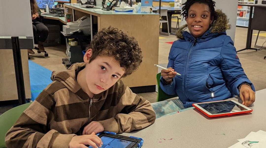 Two tweens use iPads in the library.