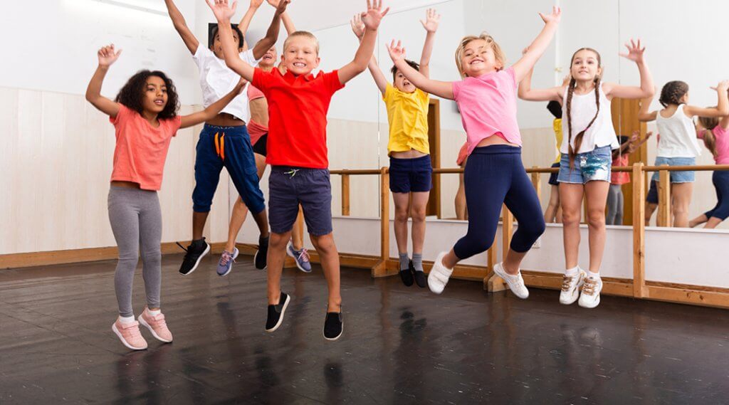 Seven elementary school age children jump with their arms raised in a dance studio.