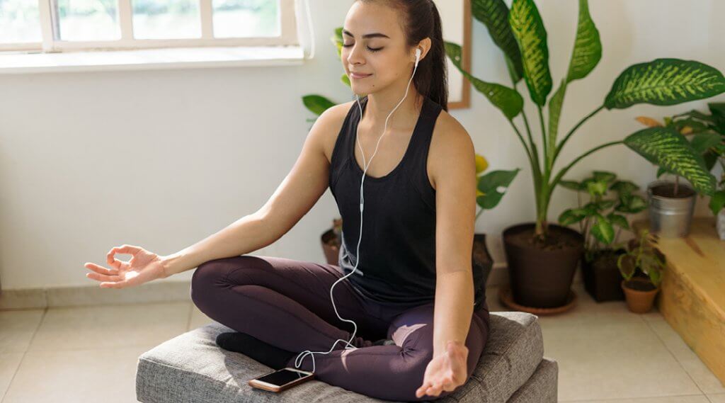 A young adult wearing earbuds sits in a meditation pose with their eyes closed.