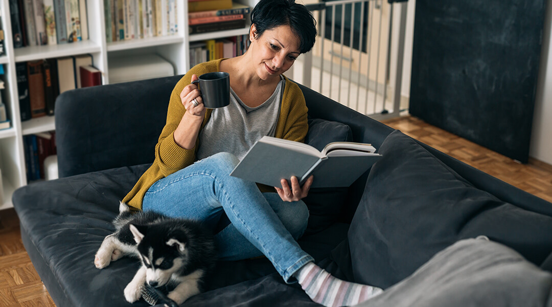 An adult sitting on sofa with their dog and reading a book.