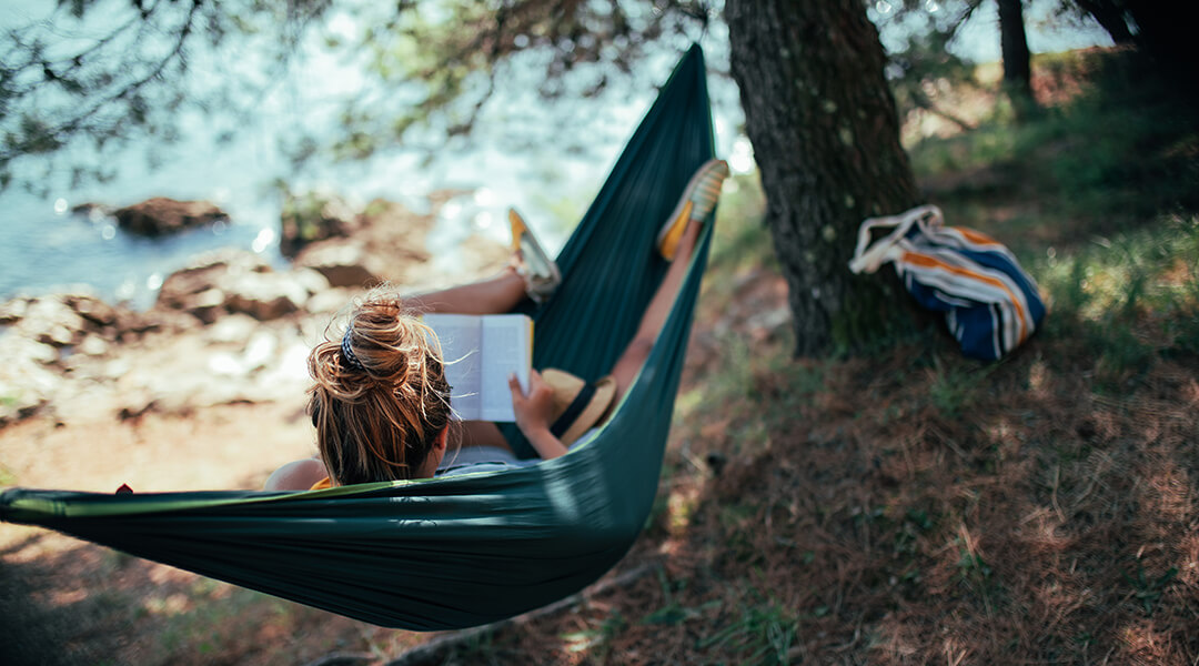 A young adult is reading a book while relaxing in hammock spread by the coastline.