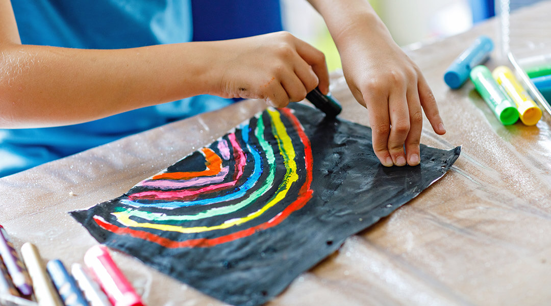 Closeup of child painting rainbow picture with different stick colors on black background.