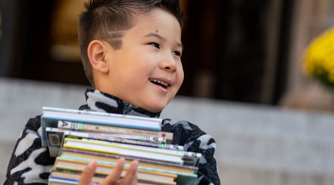 Little boy with a big smile, carrying a stack of books