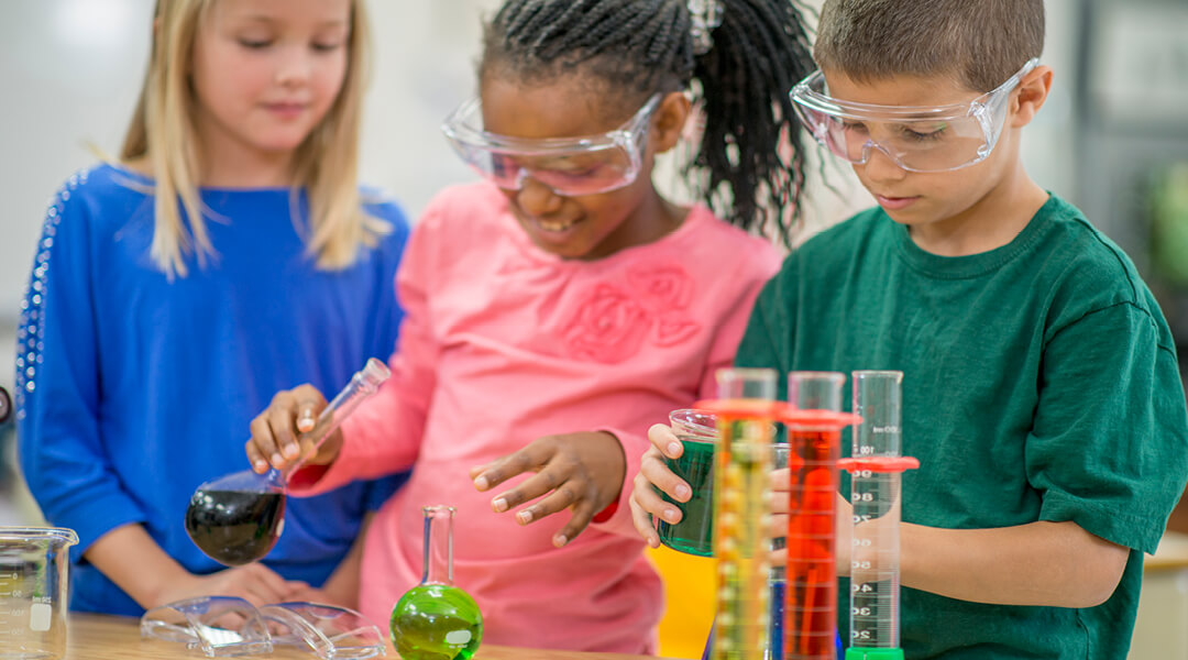 Three school-age children do a simple chemistry experiment with test tubes holding different colored liquids.