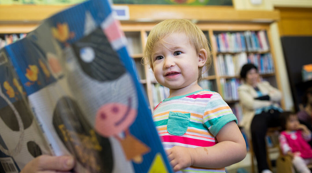 A toddler stands in front of a picture book with a cow on the cover, which is being read during storytime.
