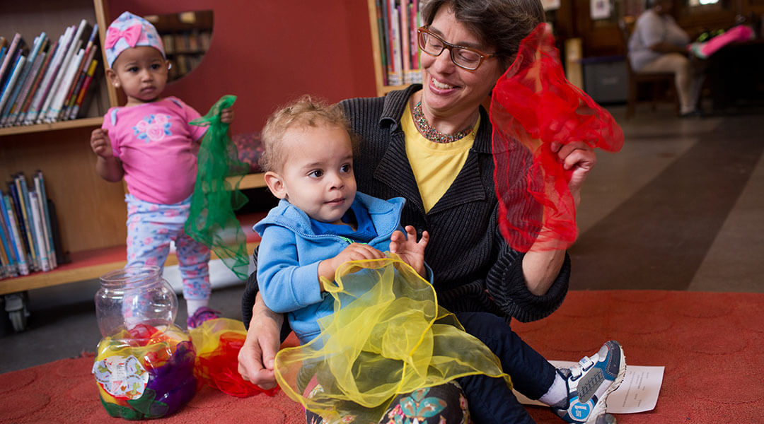 A young child sits on a caregiver's lap as they play with scarves during storytime.