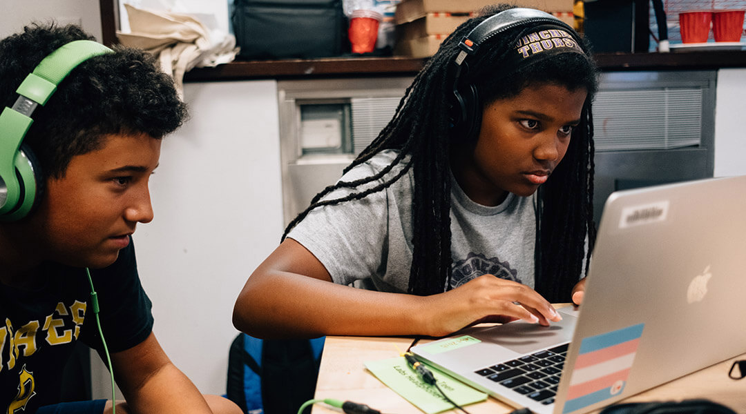 Two teens wearing headphones at a laptop.