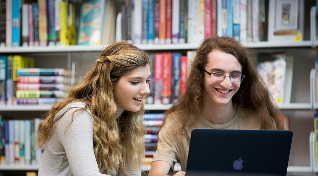 Two teenagers smile at something on a shared laptop screen.