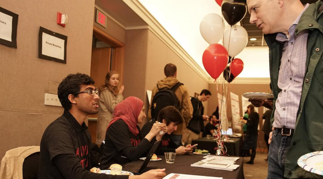 Photo of a person sitting at a table talking to another person standing across from them. Balloons and other people tabling in the background.