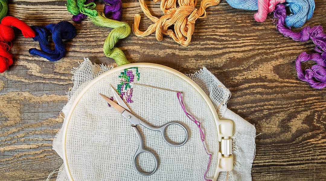 Close-up of a cross-stitch project in progress with canvas, hoop, scissors, thread and mouline on a wooden background.