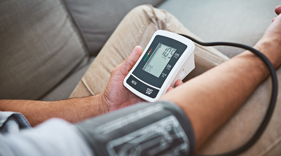 Closeup of an unrecognizable adult doing a reading of their blood pressure with a blood pressure monitor while being seated on a couch at home.